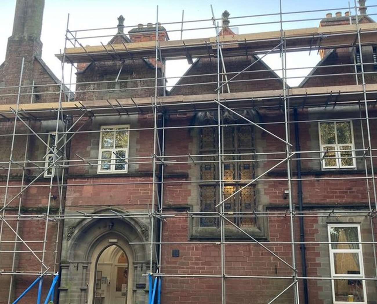 Historic brick building under restoration, covered in scaffolding. Gothic arched entrance and stained-glass windows hint at architectural elegance.