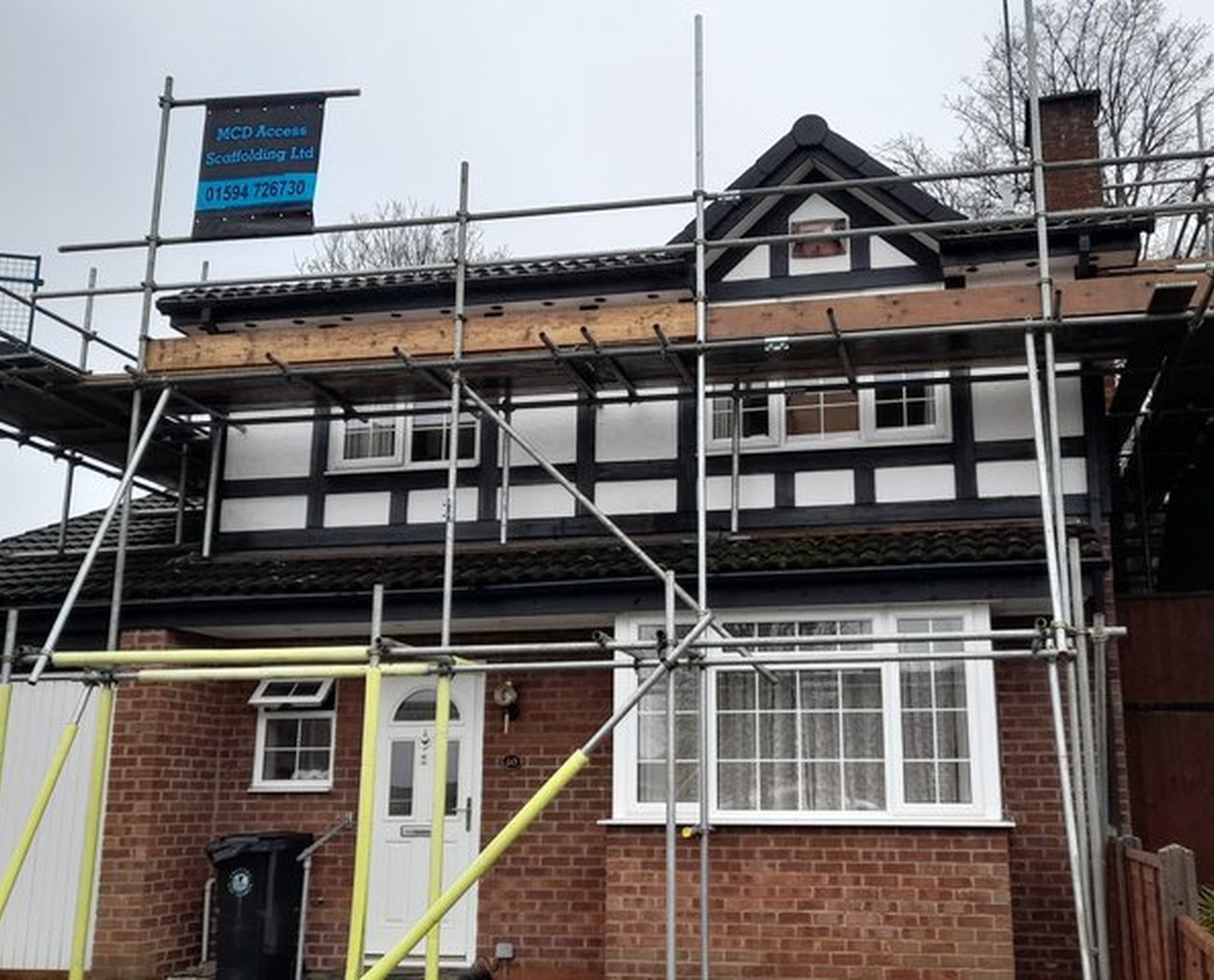 A two-story brick house with black and white timber detailing is surrounded by scaffolding, indicating repairs. The sky is overcast, creating a neutral tone.