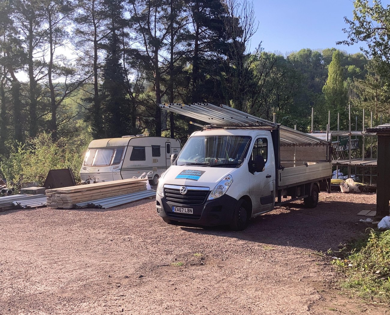 Stacks of metal scaffolding pipes and bags on a gravel surface, surrounded by trees. The setting appears outdoors, with a rustic, industrial feel.