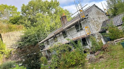 Old stone house under renovation, surrounded by scaffolding and overgrown greenery. A peaceful, rustic setting with lush trees in the background.