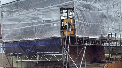 A house under construction is covered in white scaffolding sheeting. Partial view of a brick wall and window on the right, with clear skies above.